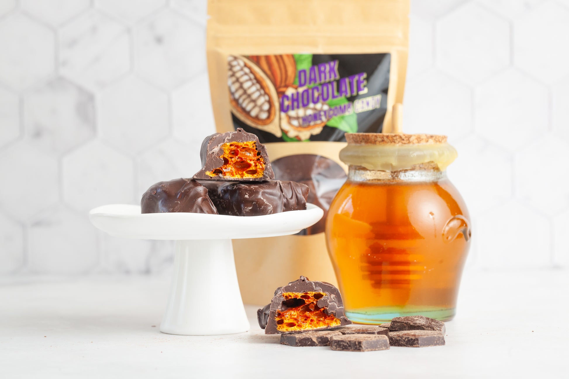 Dark chocolate covered honeycomb candy stacked on a plate. Some of the candy is cut in half to show the airy honeycomb center of the candy. There is a bag of the candy in the background and a glass pot of raw honey with a cork top covered in beeswax and chunks of chocolate in the foreground. 