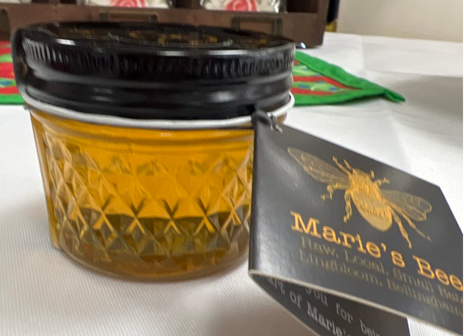 A 4oz glass jar filled with raw honey, labeled 'Marie's Bees', with a black lid and a tag attached to the jar showcasing the product details.