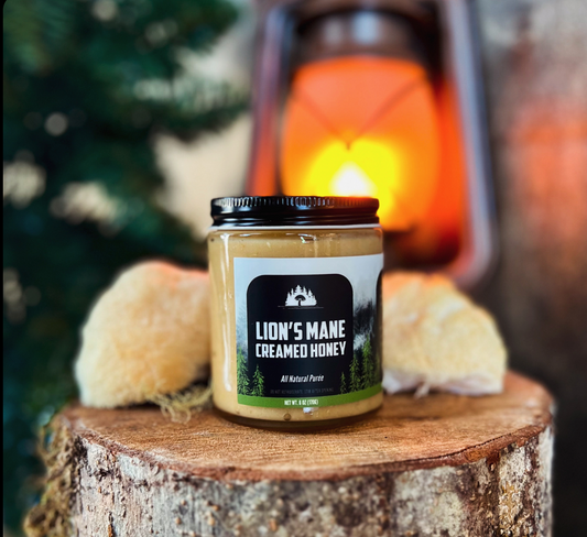A jar of Lion's Mane Creamed Honey placed on a wooden surface with a blurred background featuring a candle and evergreen trees.