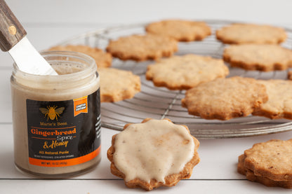 A jar of Limited Edition Gingerbread Creamed Honey by Marie's Bees, with the honey spread on cookies and a knife partially withdrawn from the jar.
