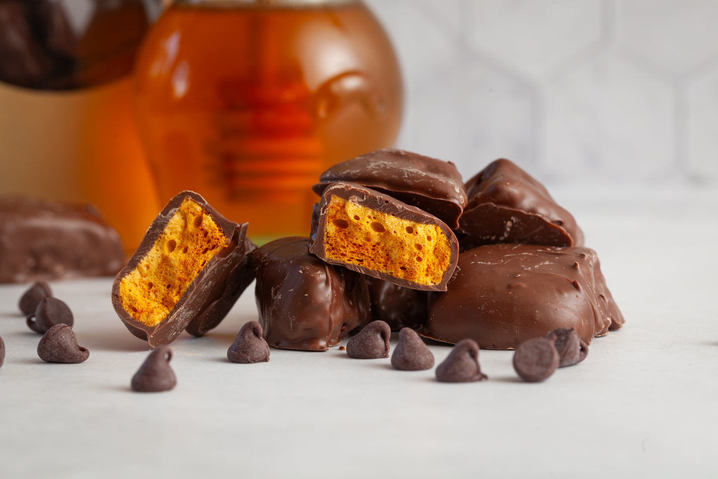 Milk Chocolate honeycomb Candy in a pile with one of the candies cut open to show the airy honeycomb center. There are chocolate chips in the foreground and a bag of candy and a glass honey pot in the background. 