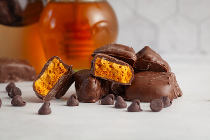 Milk Chocolate honeycomb Candy in a pile with one of the candies cut open to show the airy honeycomb center. There are chocolate chips in the foreground and a bag of candy and a glass honey pot in the background. 