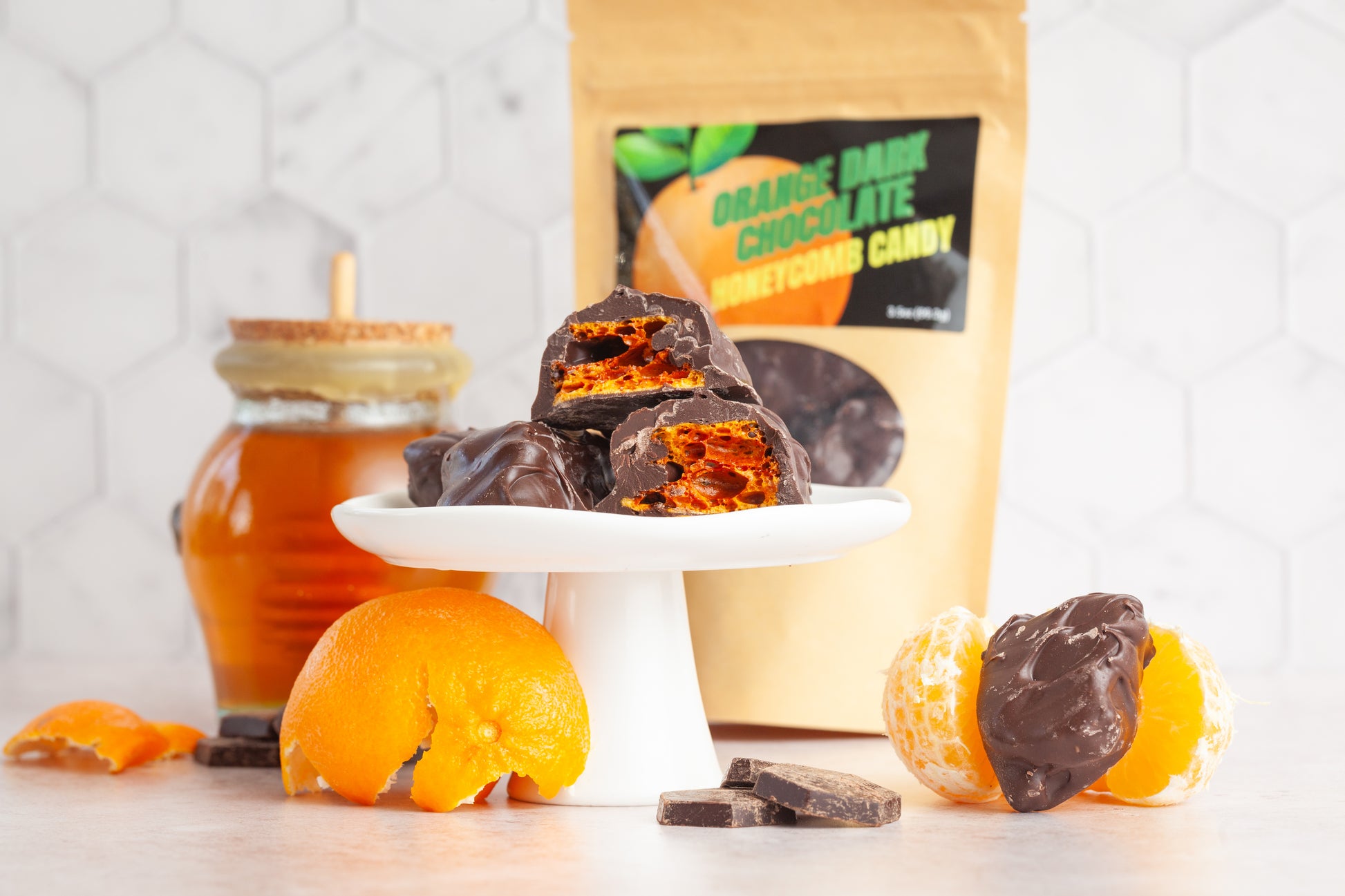 Orange Dark Chocolate honeycomb on a platter in front of a bag of the candy and a glass honeypot filled with raw honey. In the front of the photo there are oranges and chucks of chocolate. The candy is stacked on a plate cut in half to show the airy honeycomb candy inside covered with dark chocolate. 
