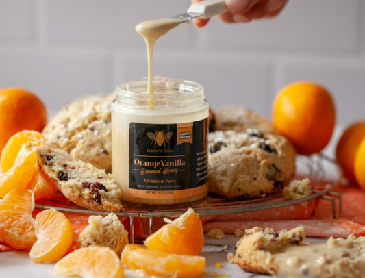 A jar of Orange Vanilla Creamed Honey with a spoonful of honey being drizzled over it, surrounded by oranges and pastries.