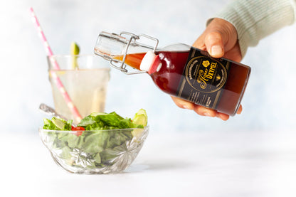 A person is pouring a dark amber liquid from a labeled glass bottle into a clear glass that contains ice and a slice of lime. A salad with dressing is visible in the background.