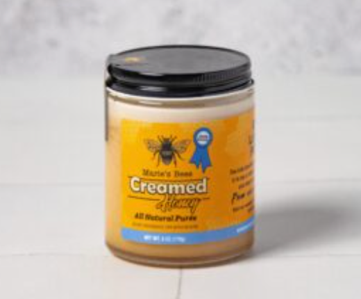 Glass jar with a black lid filled with creamed honey. The front of the label is yellow, blue and black and says Marie's Bees Creamed Honey, All natural puree 10oz