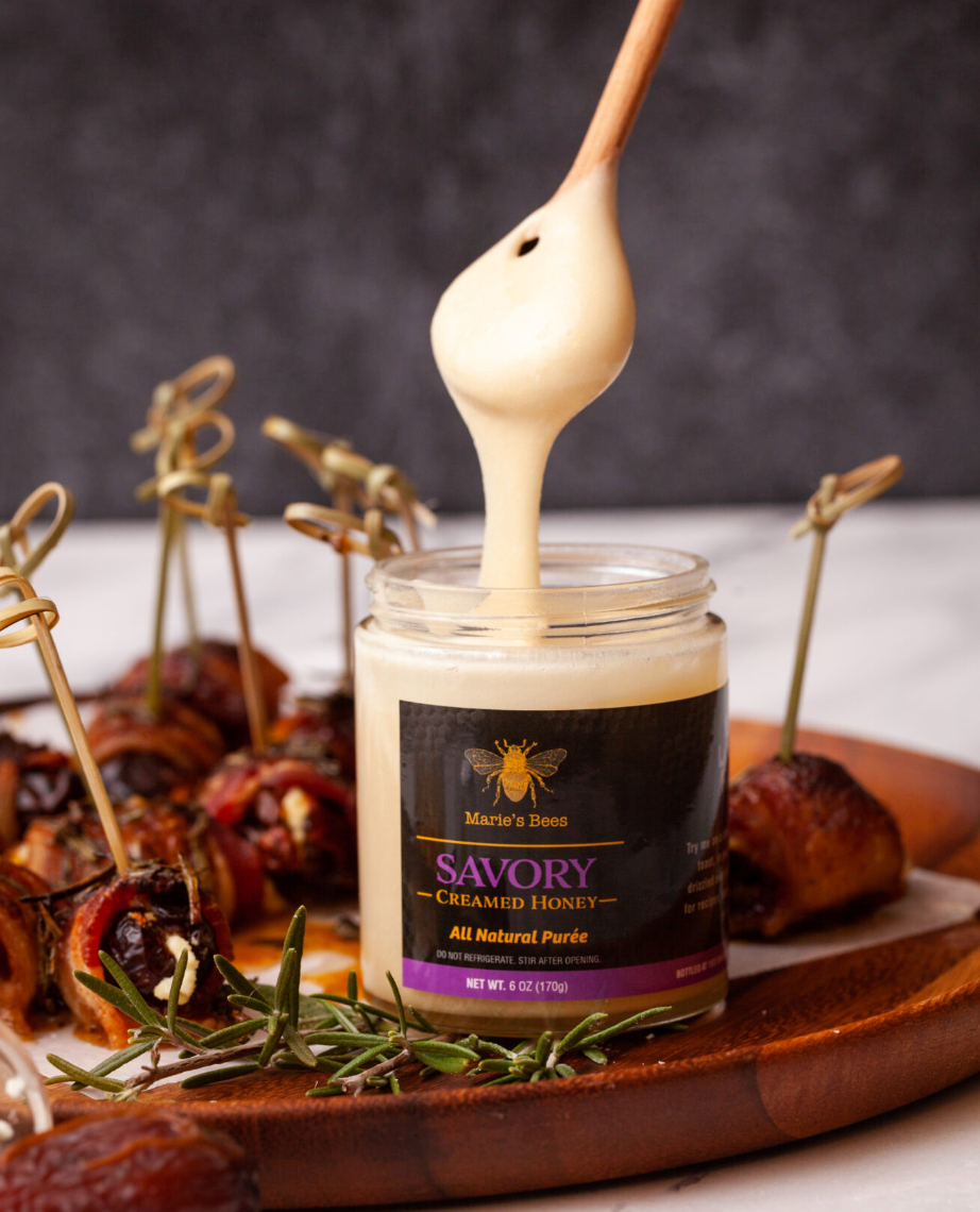 A jar of savory creamed honey with a wooden honey dipper surrounded by bacon wrapped goat cheese stuffed figs. The label reads 'Savory' and 'All Natural Honey'.