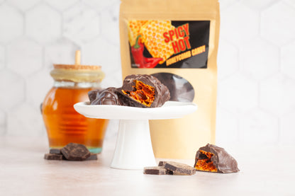 Spicy Hot Honeycomb Candy covered in chocolate. It is pictured on a platter cut up showing the candy airy honeycomb covered in chocolate. A bag of candy and a big glass jar of honey are also in the photo. 